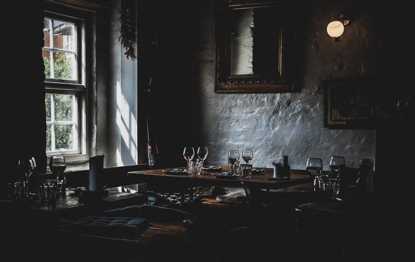 A low stone-walled room at dusk with a laid wooden table, glasses catching the light from a small window